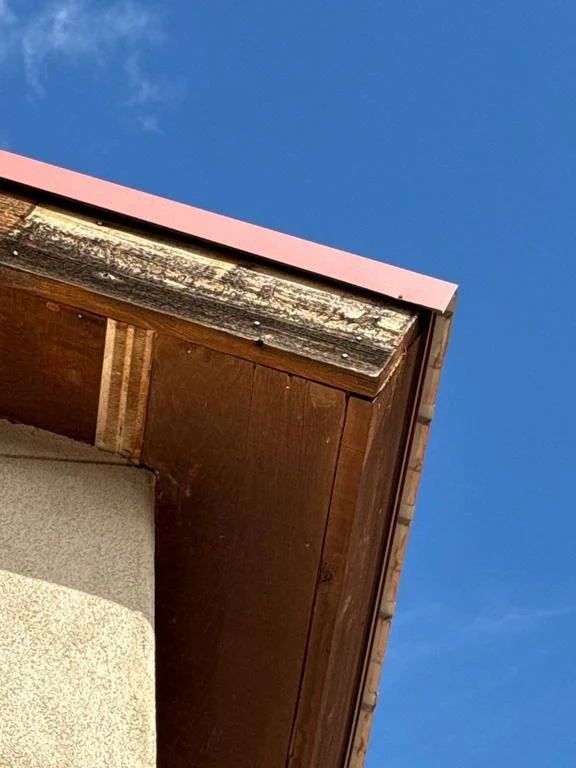 Weathered rough-cut wood fascia with peeling finish under a red metal roof