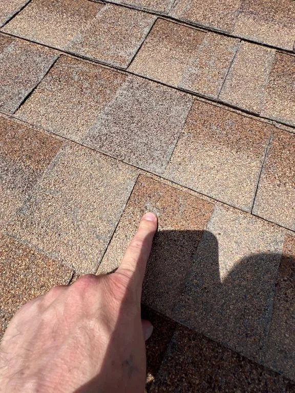 Finger pointing to a hail hit on a tan asphalt shingle